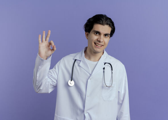 好的高兴的年轻男医生穿着医用长袍和听诊器做ok标志隔离在紫色的墙上和复印空间听诊器长袍医生