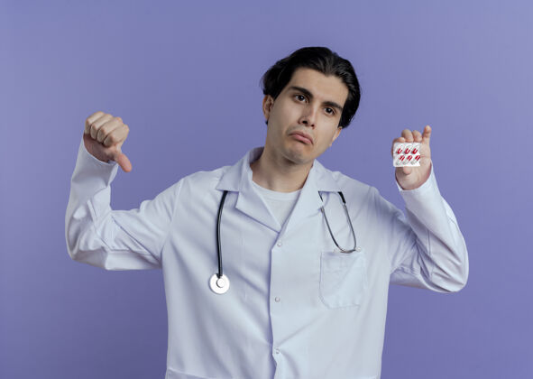 男性穿着医用长袍 戴着听诊器的年轻男医生正在一旁看着一包医用胶囊 拇指朝下被隔离在紫色的墙上向下长袍包