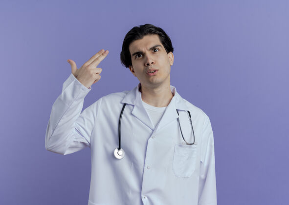 年轻令人印象深刻的年轻男医生穿着医用长袍和听诊器抬头做手势隔离在紫色墙上的复制空间手势穿