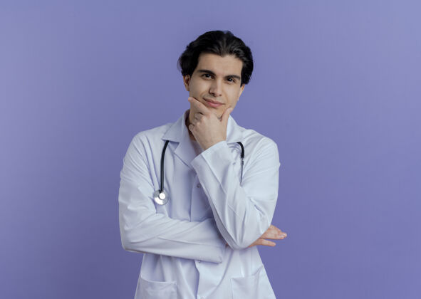 下巴高兴的年轻男医生穿着医用长袍 听诊器把手放在下巴上 隔离在紫色的墙上 留有复印空间男性长袍医生