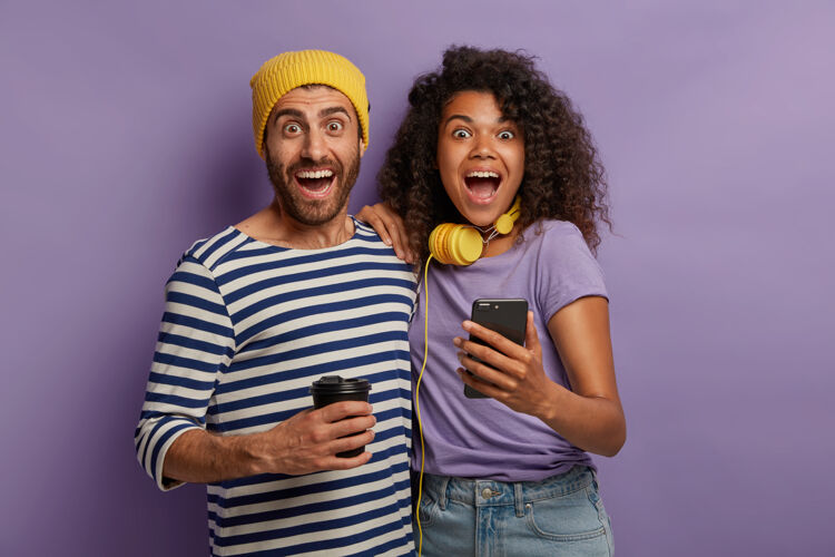 情侣快乐多样的情侣在一起的横向镜头 看社交网络上有趣的视频 拥抱和张大嘴巴 兴奋的可怕的相关性 使用智能手机 喝咖啡帽子科技小玩意