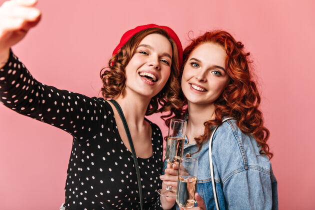 庆典美丽的女孩微笑着喝着香槟摄影棚拍摄的可爱的年轻女士拿着粉红色背景的酒杯两个人姜卷发