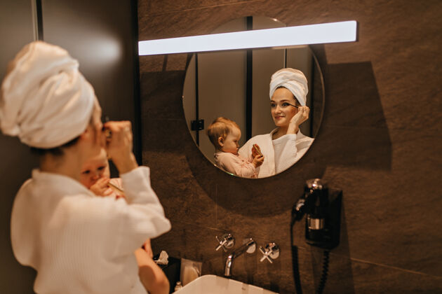 肖像穿着浴衣的迷人女人化妆 抱着孩子妈妈和女儿在浴室里观察早晨的例行公事早晨例行睫毛膏护理