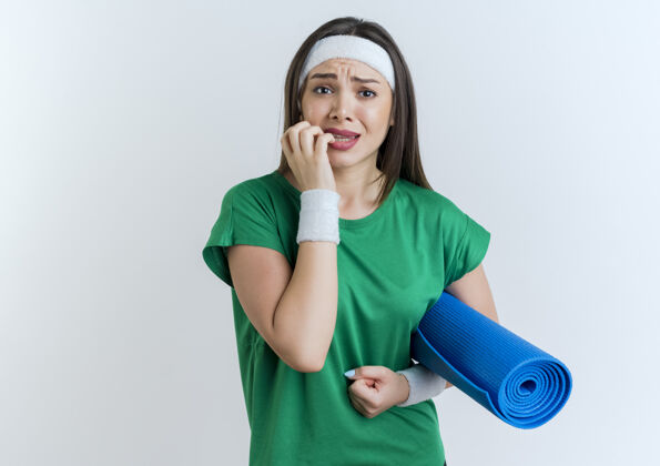 腕带焦急的年轻运动女性戴着头带和腕带看着拿着瑜伽垫咬手指焦虑运动瑜伽