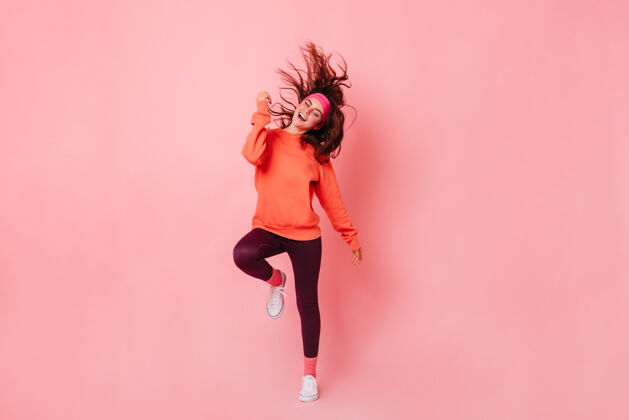 运动服穿着鲜艳运动衫和棕色紧身裤的女士在粉红色的墙上跳舞80后风格连体衣紧身裤