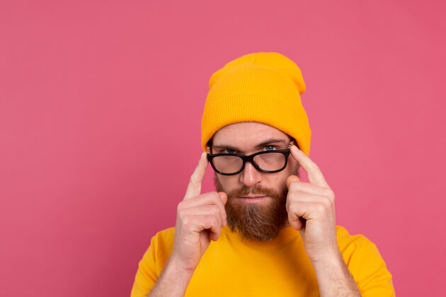 长相时尚帅气的留着胡子的欧洲男人的肖像 穿着休闲的黄色衬衫 戴着粉红色的帽子和眼镜正面站立眼镜