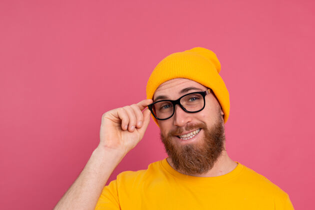 眼镜时尚帅气的留着胡子的欧洲男人的肖像 穿着休闲的黄色衬衫 戴着粉红色的帽子和眼镜站立思考墙
