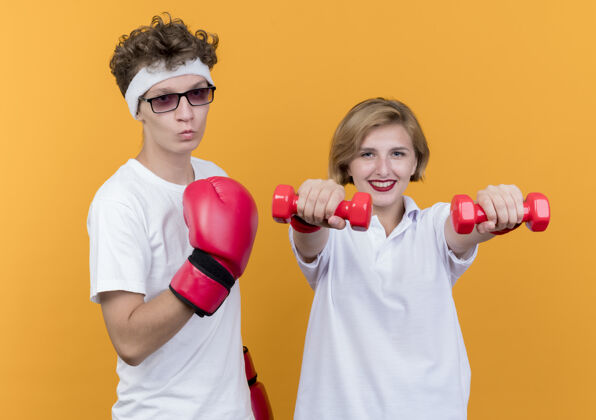 拳击一对年轻的运动型情侣 一个戴着拳击手套的女子拿着哑铃站在她的男朋友旁边 摆着橙色的姿势看站年轻