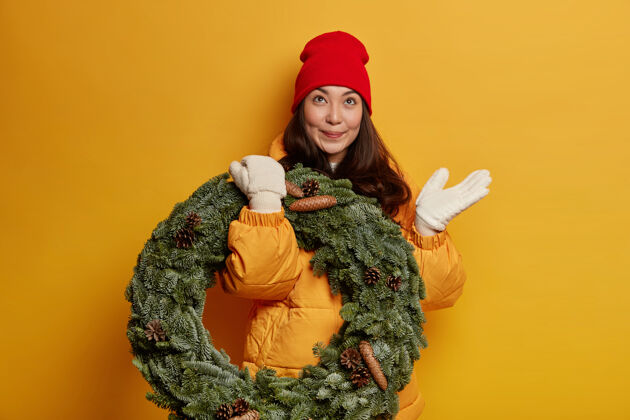 花环快乐美丽的民族妇女若有所思地看着上面 戴着红帽子 暖和的外套和白色的手套 端着绿云杉花环 想着上面庆祝圣诞节快乐携带室内