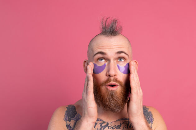 胶原蛋白欧洲帅哥留着胡子纹身上身的男人戴着紫色眼罩对着镜头摆粉红色的姿势化妆品强壮空虚
