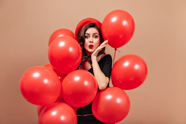 米色背景穿着红色贝雷帽和黑色连衣裙的优雅女孩吹着吻 手里拿着巨大的气球女士长波浪发