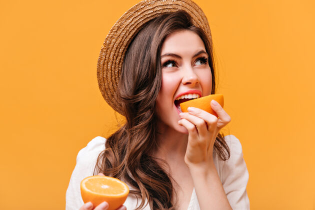 黑发戴着时髦帽子的女人咬着美味的橘子 在橘子色的背景上摆姿势波浪发微笑女人