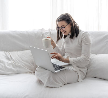 工作一个可爱的年轻女子正坐在家里的白色沙发上 穿着白色的裙子 对着笔记本电脑培训房子女人