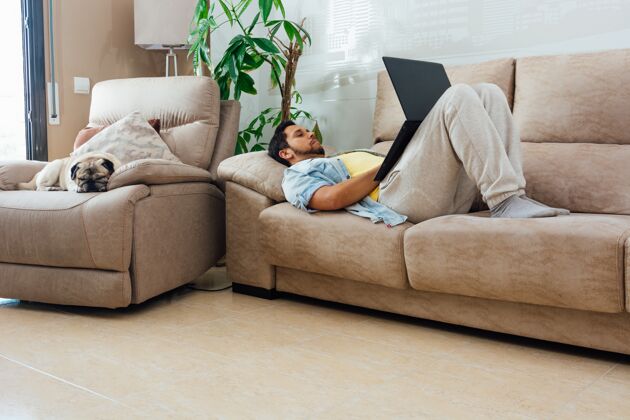男人年轻人在家里的沙发上休息 用笔记本电脑和他的狗在他身边放松成人扶手椅