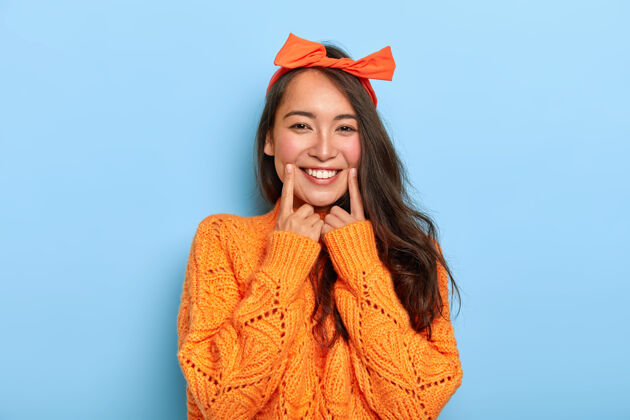 女性快乐的黑发韩国女孩的肖像 带着牙齿般的微笑 食指靠近嘴巴 戴着橙色的头带和针织毛衣服装年轻人人