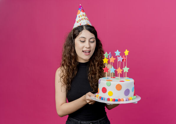 积极一个年轻漂亮的女人 卷发 戴着节日帽 手里拿着生日蛋糕 生日快乐 积极的生日派对概念 粉色的女人派对年轻