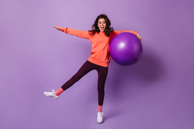 卷发快乐的卷发女运动员在紫色工作室与巨大的健身球乐趣运动衫80后身体