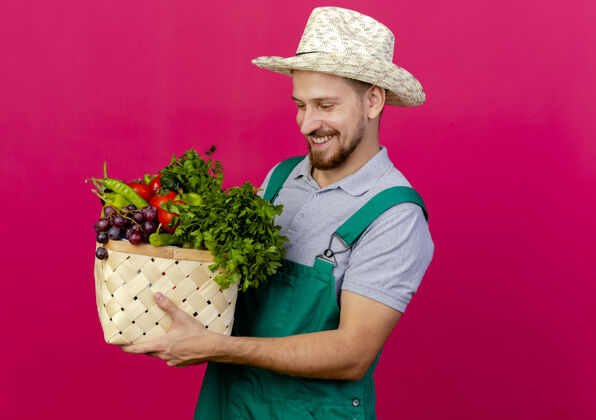 深红色面带微笑的年轻英俊的斯拉夫园丁 穿着制服 戴着帽子 看着一篮子蔬菜人制服年轻人