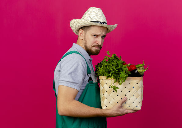 视图皱眉的年轻英俊的斯拉夫园丁 穿着制服 戴着帽子站在侧视图中 手里拿着一篮子蔬菜看男人深红色