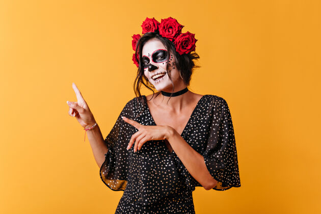 深色橙色背景上的墨西哥女孩的正面肖像 留有文字空间戴着头盖骨面具的女人可爱地笑着 手指向上脸化妆品拉丁