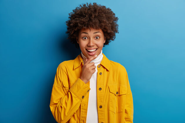 自然可爱微笑的非裔美国人 在镜头前带着惊喜的表情 听到喜讯 穿着亮黄色的夹克 在蓝色的背景下与世隔绝女人人类发型