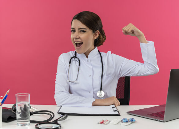医生快乐的年轻女医生穿着医用长袍和听诊器坐在办公桌前 拿着医疗工具和笔记本电脑做着坚强的手势年轻笔记本电脑女士
