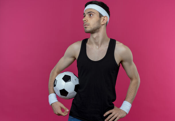 抱着戴着头巾 拿着足球 表情严肃地站在粉色墙壁上的年轻人看站着年轻