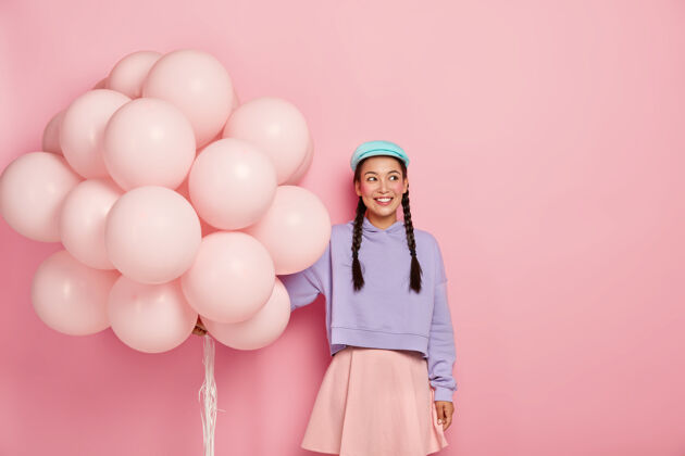 梦幻快乐满意女孩的肖像 长辫子 穿着宽松的毛衣 裙子 有最小的化妆品 站在粉红色的墙上膨胀气球派对女士帽子