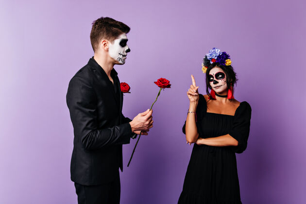 女人画着脸的女孩很不高兴 因为她的男朋友只给了她一朵玫瑰一男一女穿着黑色衣服 在淡紫色的背景上摆姿势节日文化死亡