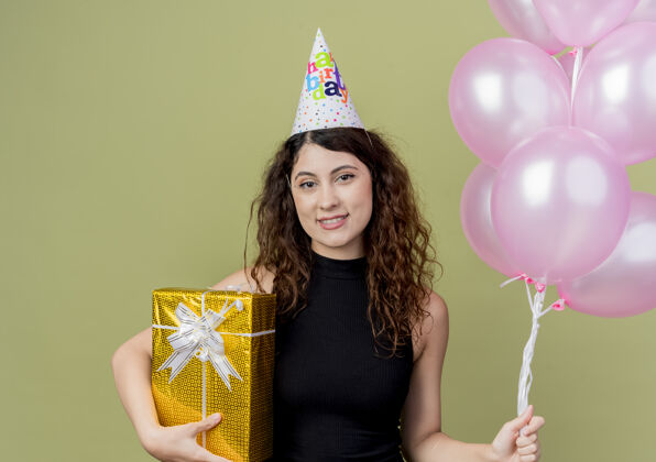 抱着一个年轻漂亮的女人 卷发 戴着节日帽 手里拿着气球和生日礼物 脸上带着幸福的笑容站在明亮的墙上空气帽子女人