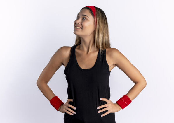 微笑身穿黑色运动服 头戴红色头巾的年轻健身女孩站在白墙上 自信地微笑着 双臂放在臀部手臂自信年轻