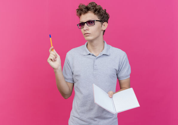 马球一个戴着黑眼镜 穿着灰色马球衫的年轻人 手里拿着一本带钢笔的笔记本 一脸严肃地站在粉红色的墙上眼镜脸站着