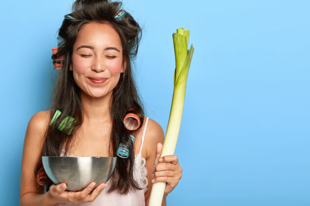 生的高兴的年轻女模特带着卷发器 端着绿韭菜和碗 做健康的沙拉 闭上眼睛室内吃蔬菜