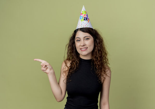 女人年轻漂亮的女士 卷发 戴着节日帽 手指指向一边 微笑着 站在轻墙上的生日派对概念轻帽子卷发