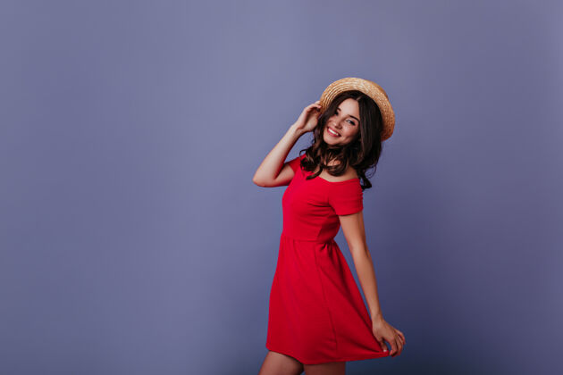 深褐色迷人的白人女孩跳舞和触摸她的夏季帽子室内肖像的微笑美丽的年轻女子穿着红色连衣裙积极休闲服饰