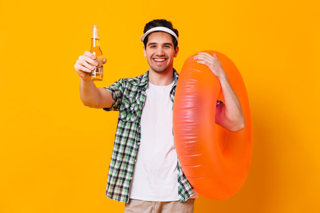 帽子一个戴着帽子 穿着衬衫和t恤的男人的肖像 他手里拿着一瓶啤酒和一个充气圈 在一个孤立的空间里潜水面具短裤男性