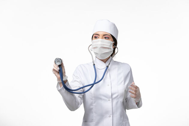 消毒正面图女医生身穿白色无菌医疗服 戴口罩因冠状病毒-使用听诊器对轻白墙病病毒冠状病毒-大流行性疾病听诊器视图套装