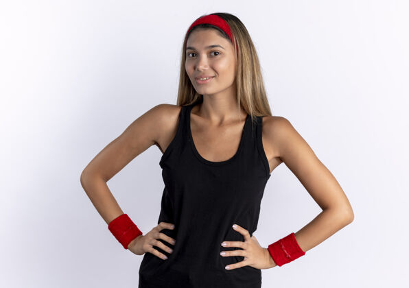 健康身穿黑色运动服 头戴红色头巾的年轻健身女孩站在白色的墙上 自信地微笑着 双臂放在臀部站立年轻头带