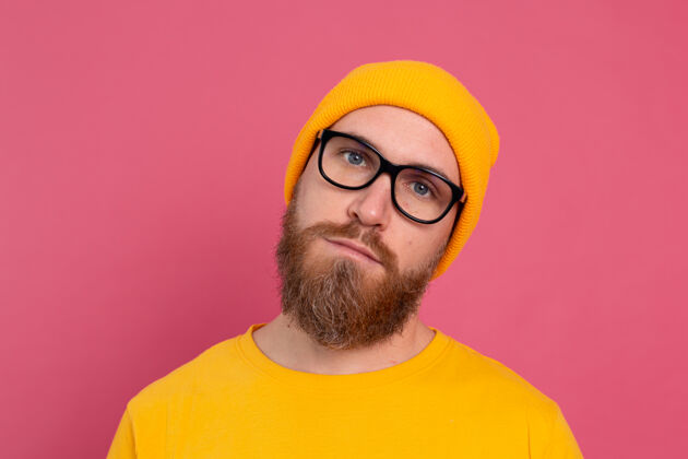 胡须时尚帅气的留着胡子的欧洲男人的肖像 穿着休闲的黄色衬衫 戴着粉色背景的帽子和眼镜眼镜严肃自信