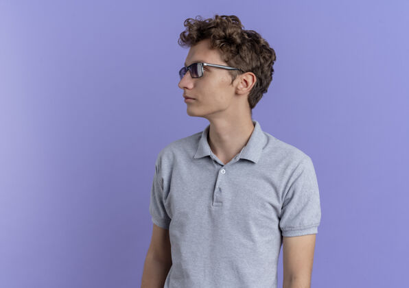 眼镜一个戴着黑眼镜 穿着灰色马球衫的年轻人站在蓝色的墙上 严肃地看着一边男人站看