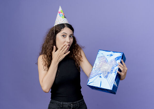 帽子一个年轻漂亮的女人 卷发 戴着节日帽 手里拿着生日礼盒 站在蓝色的墙上 看着惊喜的生日派对概念惊讶抱着站着
