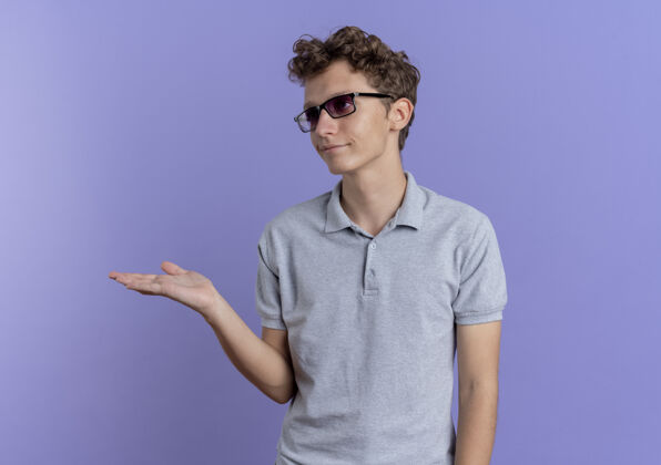 年轻一个戴着黑眼镜 身穿灰色马球衫的年轻人站在蓝色的墙壁上 用手举着什么东西 带着怀疑的表情看手臂男人