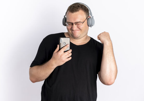 男人一个戴眼镜的超重男人 穿着黑色t恤 头戴耳机 手持智能手机 站在白墙上欣赏他最喜欢的音乐喜欢抱着眼镜