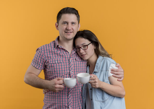 衣服一对穿着休闲服的年轻夫妇手持咖啡杯快乐而积极地站在橙色的墙上抱着年轻站着