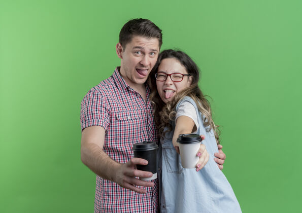 年轻一对穿着休闲服的年轻夫妇在绿色的墙上展示咖啡杯 一起玩得很开心展台女人休闲