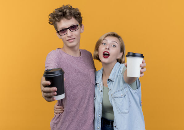 情侣一对年轻漂亮的情侣穿着休闲服展示咖啡杯微笑着站在橙色的墙上年轻人穿着表演