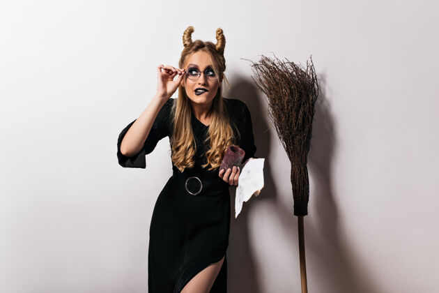 拼写戴着眼镜的优雅巫师在白墙上摆着滑稽的姿势金发女巫站在扫帚旁边庆典邪恶恐惧