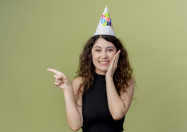轻戴着节日帽的卷发的年轻漂亮女人微笑着 用手指着一面生日派对的概念站在光墙上站侧线卷发
