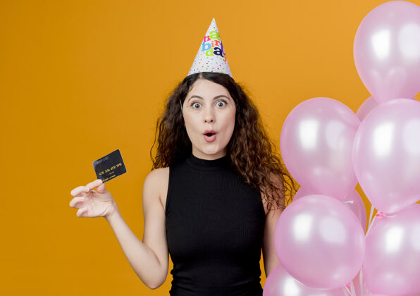 女人一个年轻漂亮的女人 一头卷发 戴着节日帽 手里拿着气球 手里拿着信用卡 站在橙色的墙上 惊讶地看着生日派对的概念惊喜立场帽子