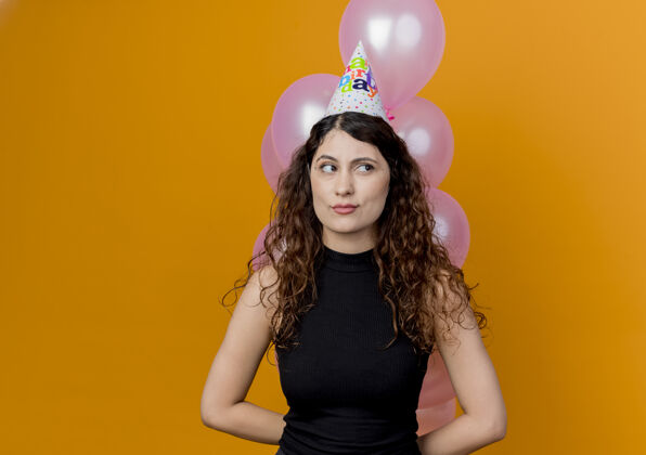 一边一个年轻漂亮的女人 卷发 手持一束气球 带着怀疑的表情站在橙色的墙上 看着旁边的生日派对看着女人站着